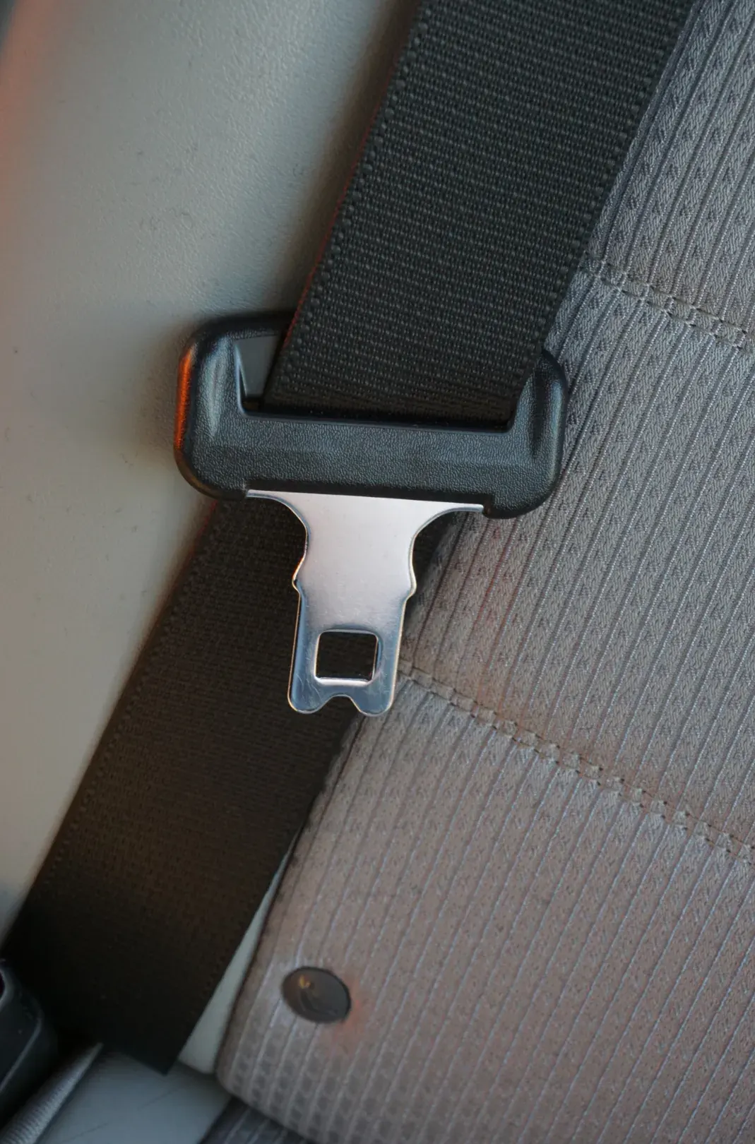 Seat belt webbing running through a moulded guide into a steel latch plate, photographed against a light vehicle interior trim panel